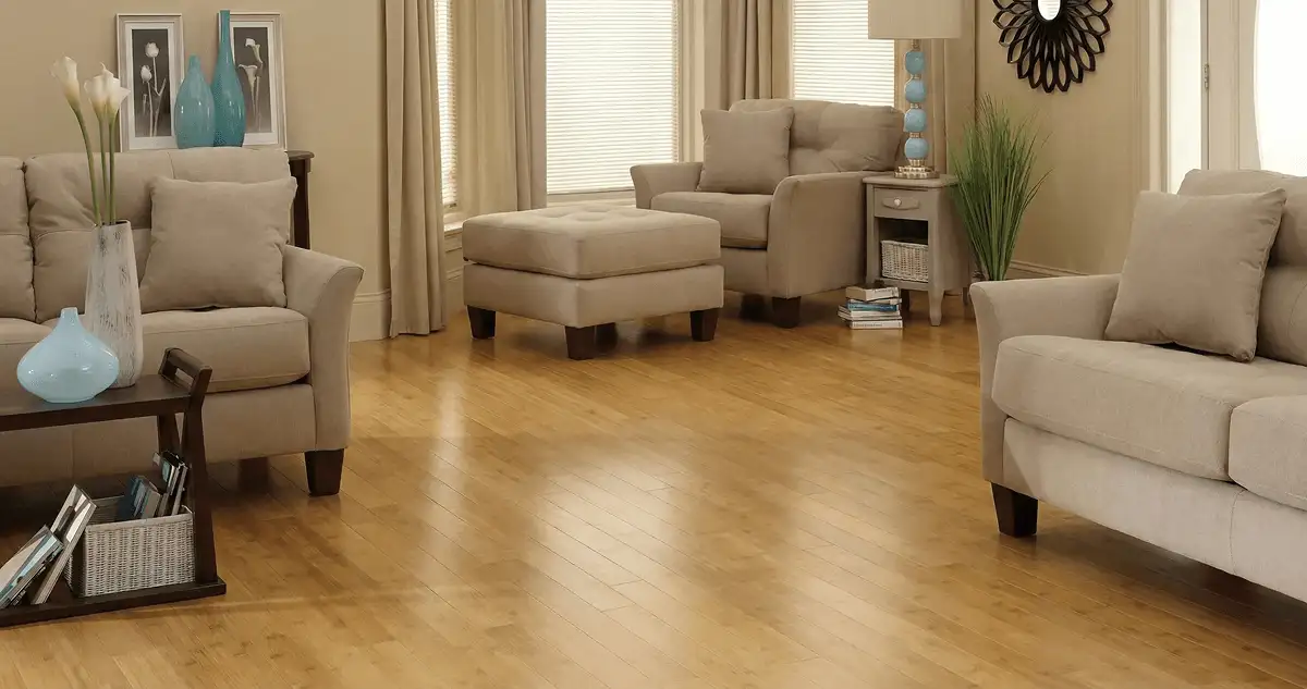 A living room with professionally installed bamboo flooring, beige sofas, a matching ottoman, a side table with a blue lamp, vases, framed pictures, and large windows with blinds and curtains in New Orleans, LA